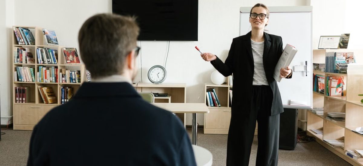 A teacher in a black blazer conducts a session with a student in a library setting.
