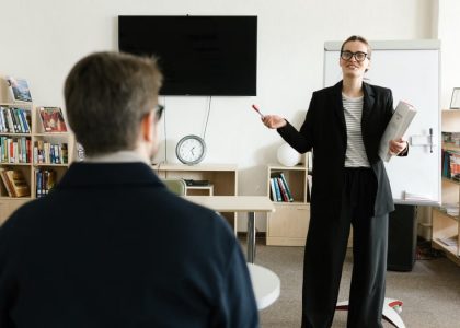 A teacher in a black blazer conducts a session with a student in a library setting.